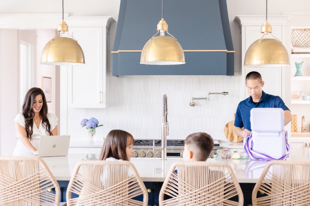 Monica working on a laptop while Michael packs lunches with their kids in the kitchen