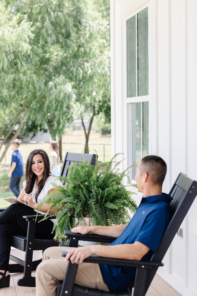 Monica taking notes on the porch while Michael sits nearby and the kids play in the yard